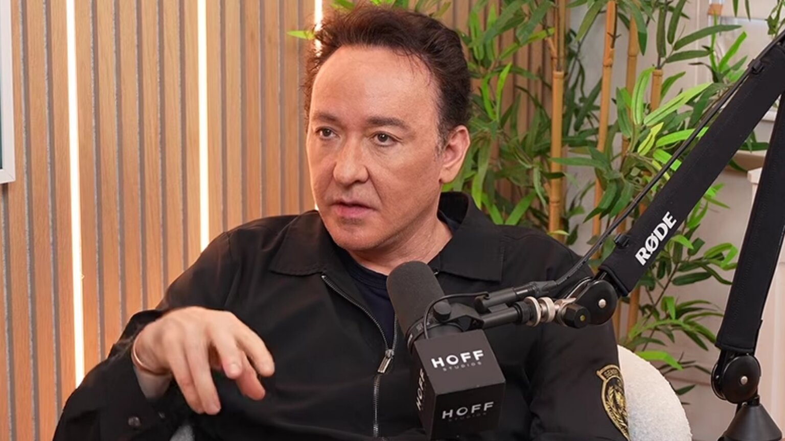 John Cusack speaks during a podcast interview, gesturing beside a studio microphone.
