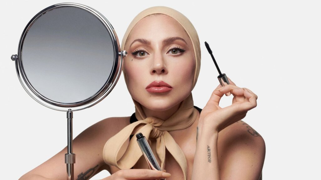 Lady Gaga holding a Haus Labs mascara wand beside a round makeup mirror.