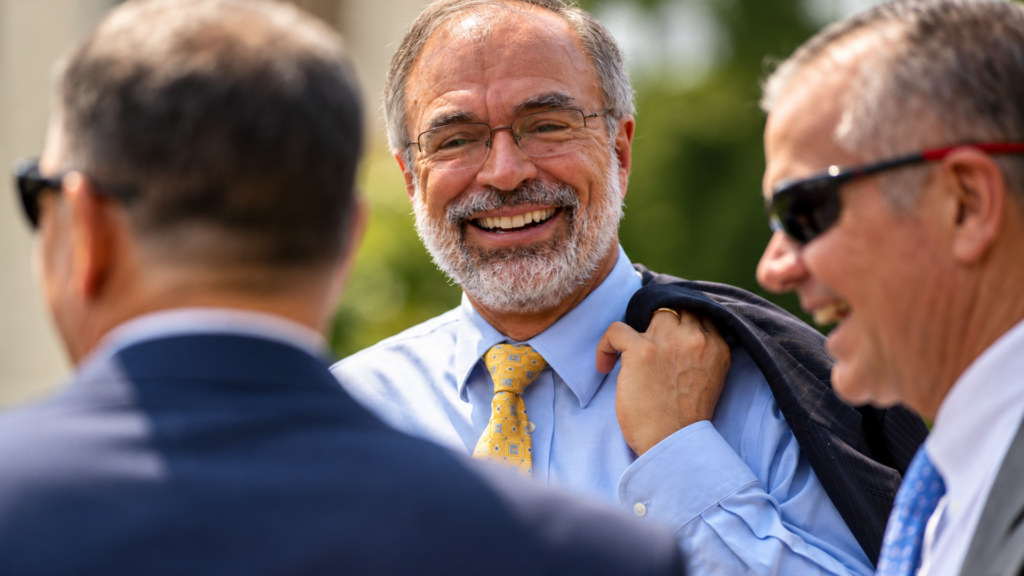 Smiling man in a suit with glasses talking outdoors with colleagues while holding his jacket over his shoulder.