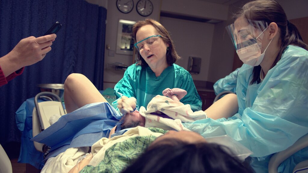 Medical professionals wearing protective gowns and face shields assist a woman during childbirth in a hospital delivery room, with a newborn baby visible.