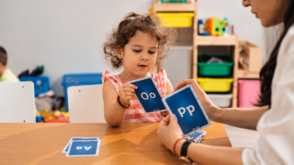 A young child sitting at a table learning letters from flashcards held by an adult.