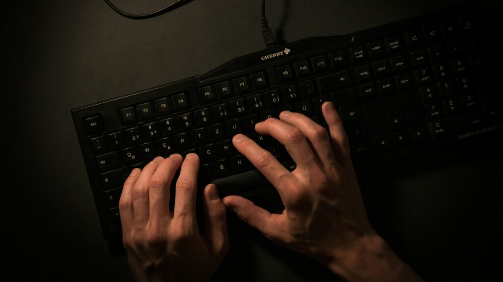 Hands typing on a black computer keyboard in a dimly lit setting.