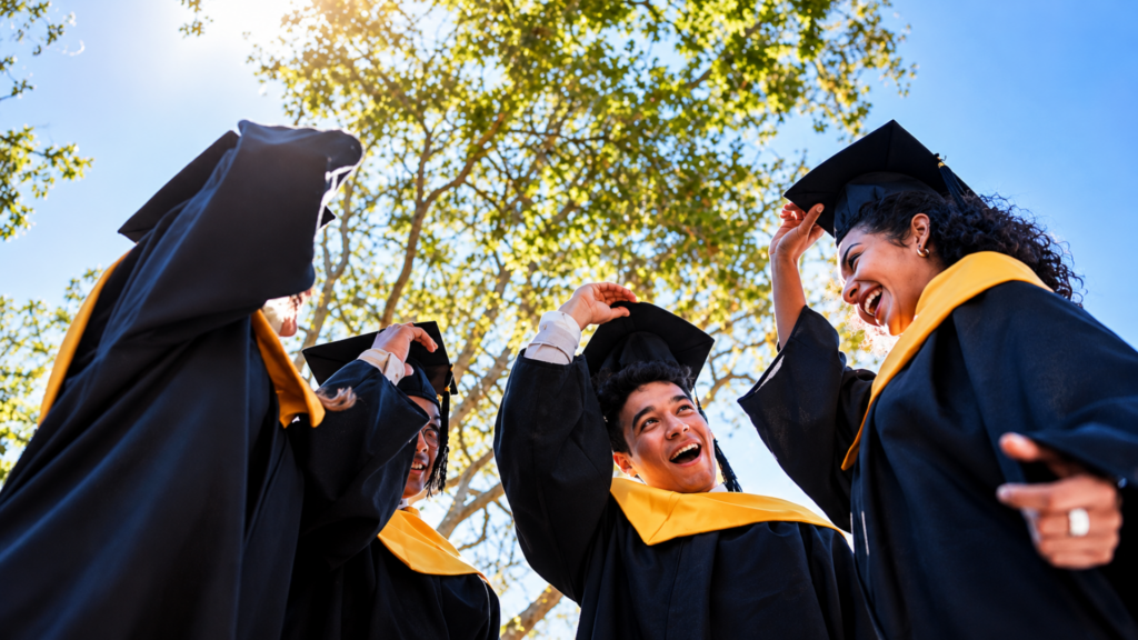 Low-angle shot of joyful graduates in cap and gown smiling together outdoors under a tree.
