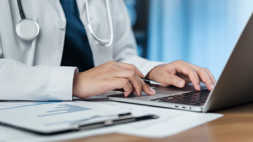 Doctor typing on a laptop with medical charts on a desk.