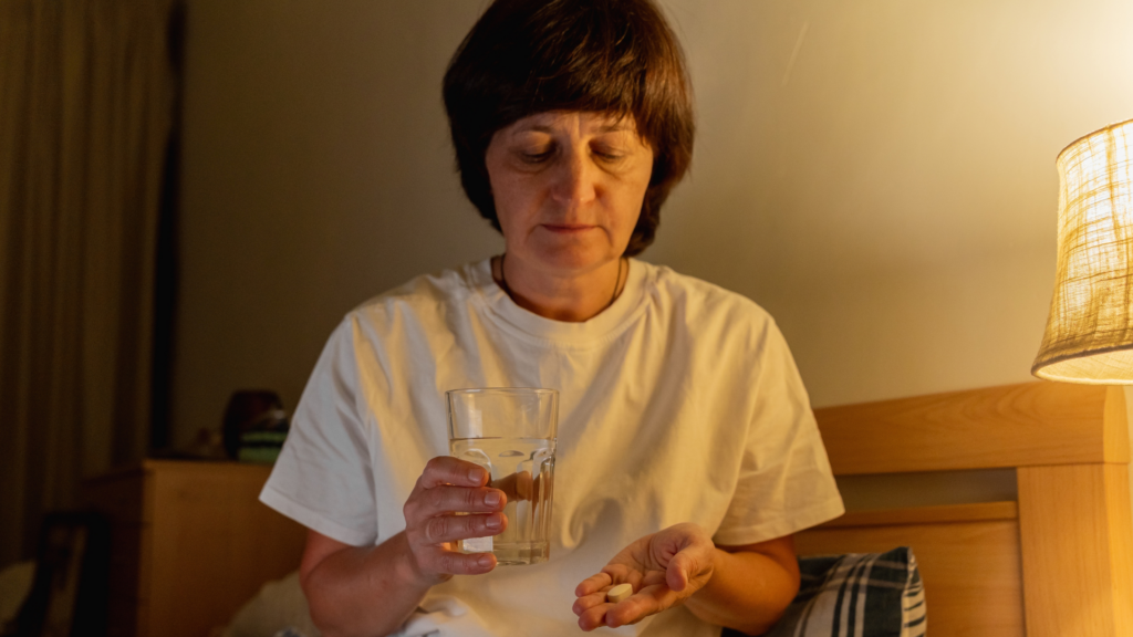 Woman holding a glass of water and pill by a bedside lamp.