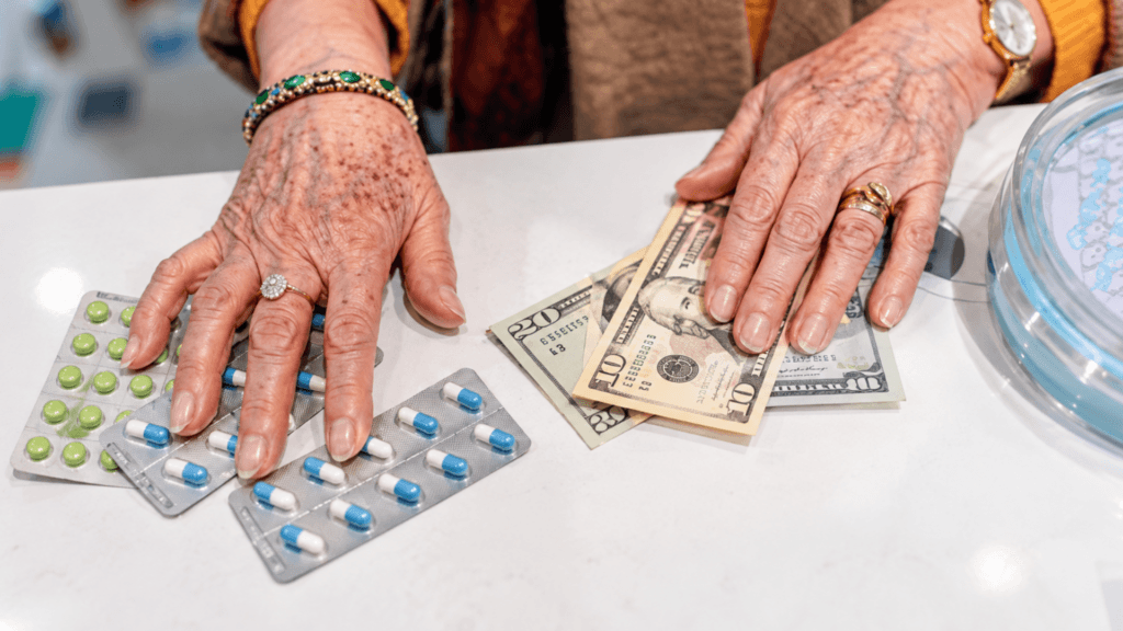 An elderly person's hands resting on blister packs of medication and US dollar bills on a counter.