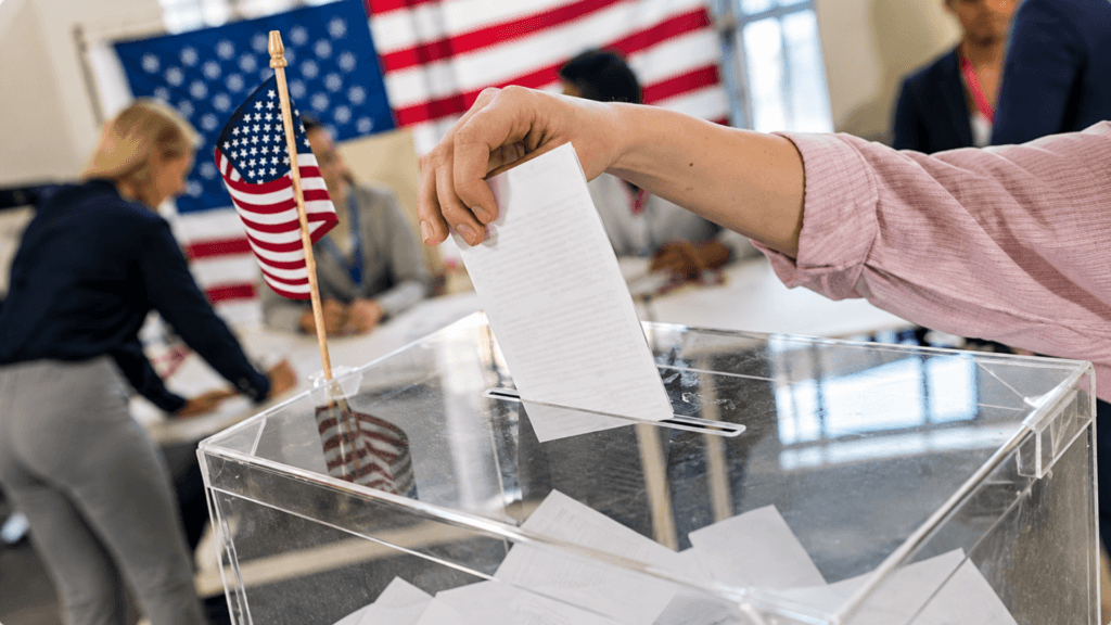 A close-up of a person dropping a paper ballot into a clear plastic ballot box next to a small American flag.