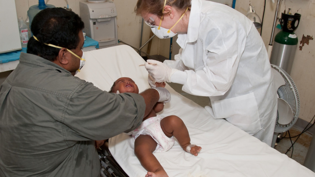 Doctor in protective gear treating a baby on a hospital bed with a parent present.