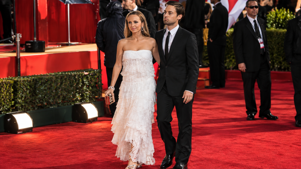 Tobey Maguire in a black suit walking the red carpet with Jennifer Meyer wearing a tiered white lace dress.