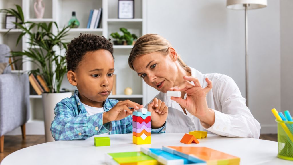 Child therapist or educator guiding a young boy as he carefully stacks colorful wooden blocks at a table.