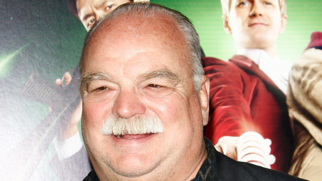 Close-up of Richard Riehle smiling in front of a movie poster.