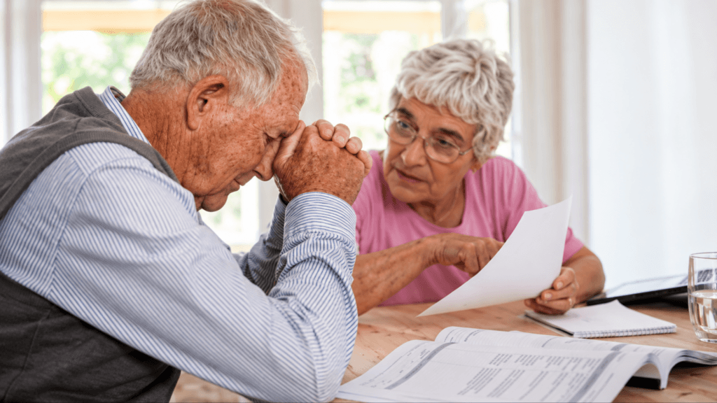 An elderly man sitting with his head resting in his hands in distress while a woman beside him points at a document on a table.