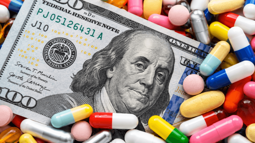 A US hundred-dollar bill partially covered by a scatter of colorful prescription pills and capsules.