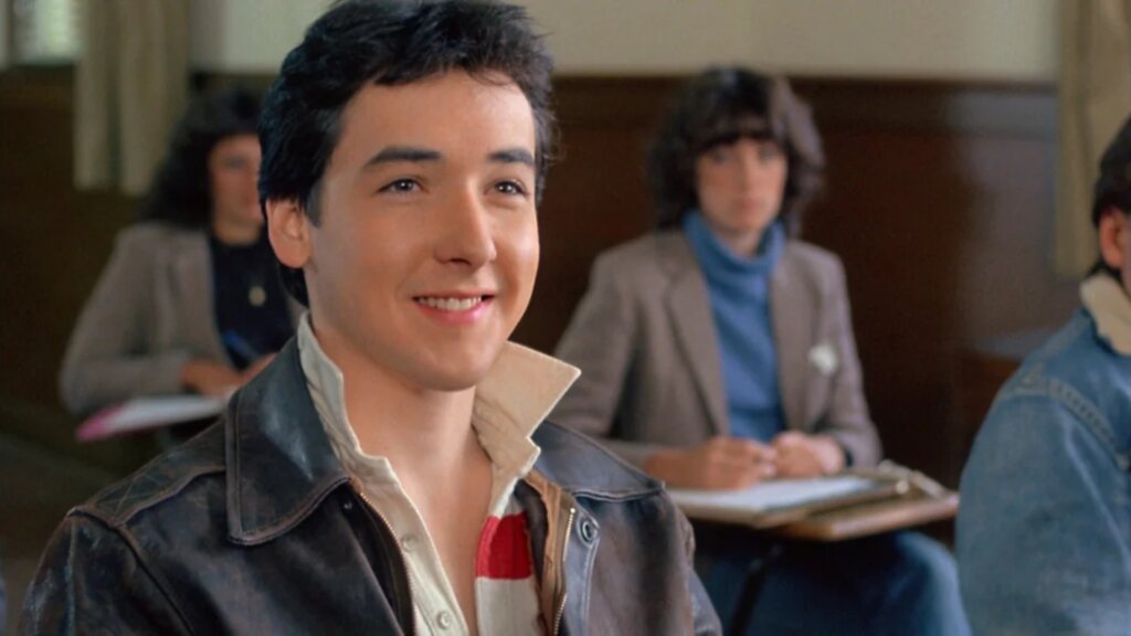 A young John Cusack smiles while sitting in a classroom scene from The Sure Thing.