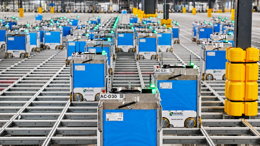 An automated Ocado grid system with multiple blue robotic bots operating across a warehouse floor.