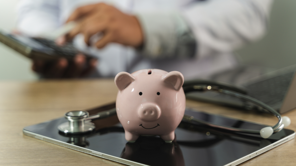 A piggy bank and stethoscope resting on a tablet, with a person calculating in the background.