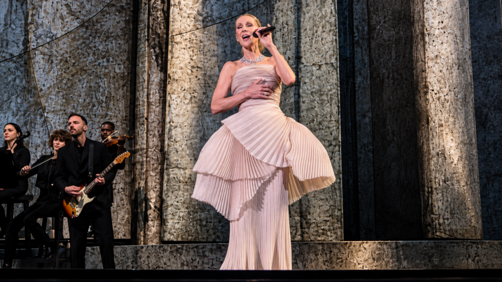 Celine Dion singing on stage wearing a blush pink pleated gown accompanied by live musicians.