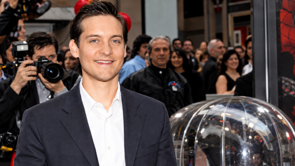 Actor Tobey Maguire smiling in a suit while surrounded by photographers on a red carpet.