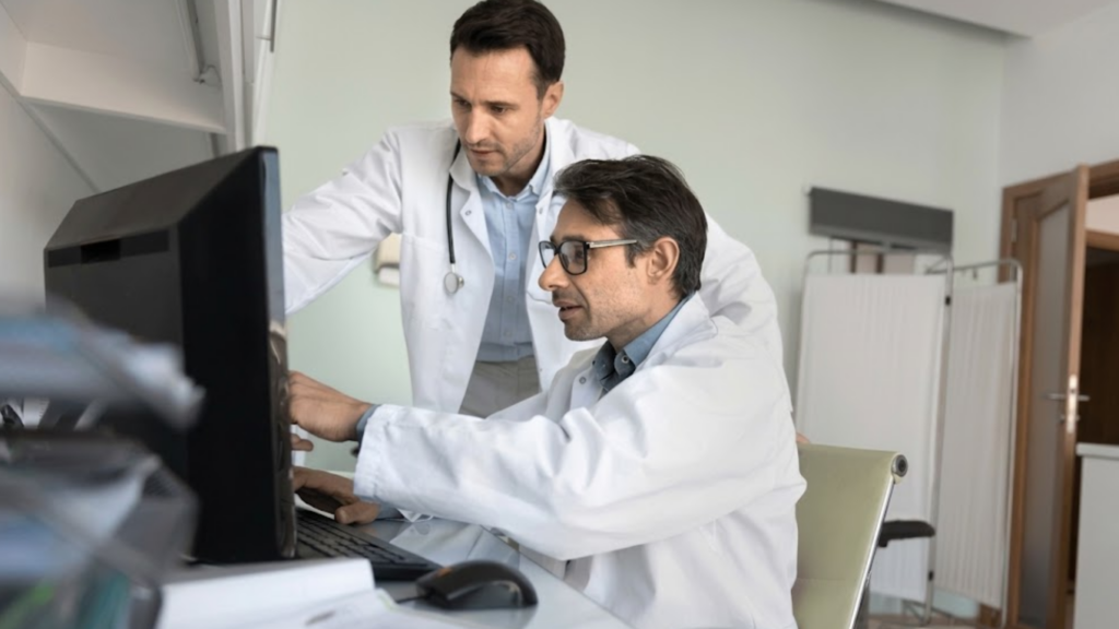 Teamwork in medicine. Two male doctors talk by computer screen at the hospital office discussing difficult patient cases, brainstorming diagnoses and analyzing medical records.