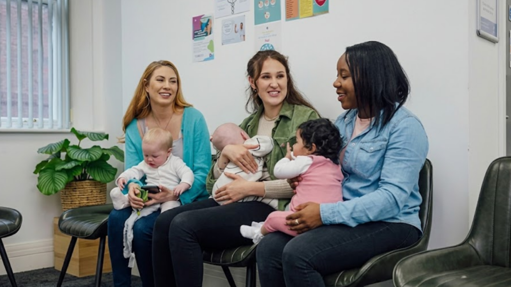 Three women sit in a waiting room, each holding a baby, engaged in conversation while waiting.
