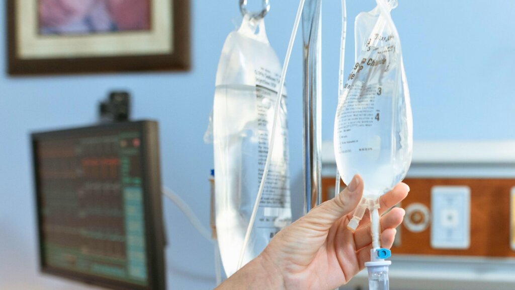 Healthcare worker checking intravenous fluid bags connected to an IV pump in a hospital treatment room.