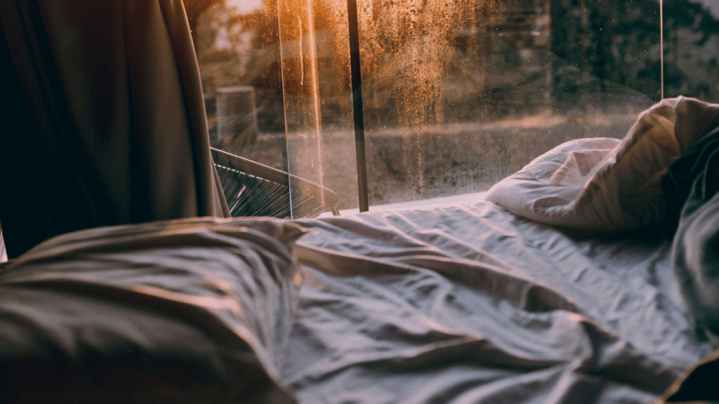 A cozy bed with rumpled linens positioned next to a large glass window, capturing the warm glow of sunrise over trees outside.