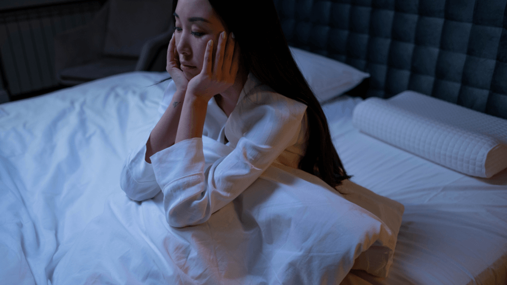 A woman sitting on a bed resting her head on her hands, appearing pensive or troubled in dim blue night lighting.