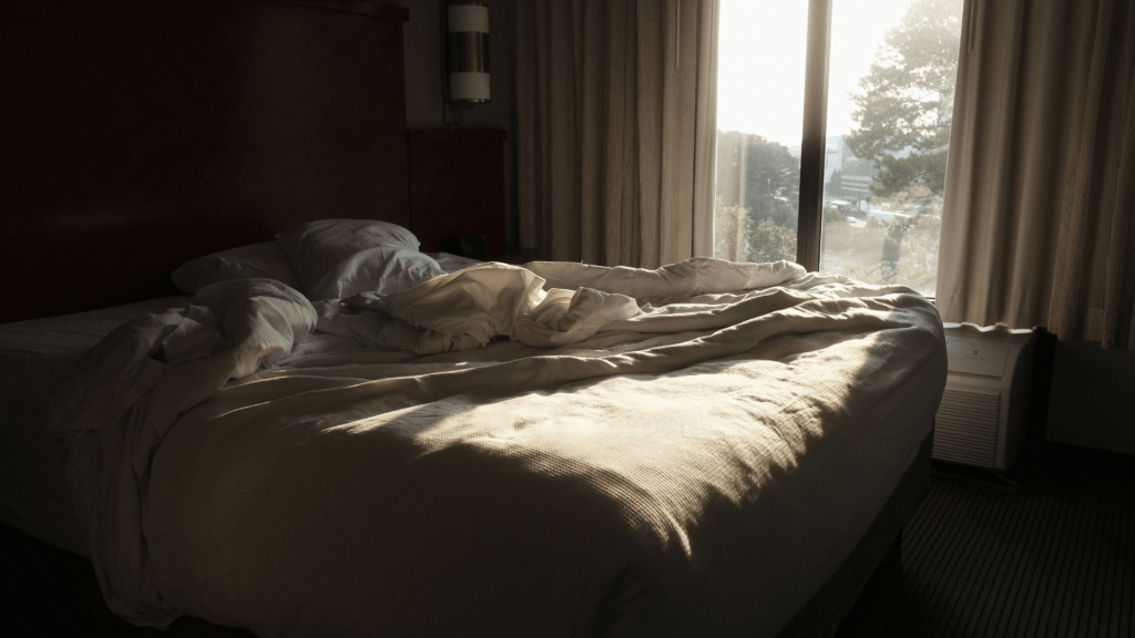 An unmade hotel bed illuminated by natural daylight streaming in from an adjacent window.