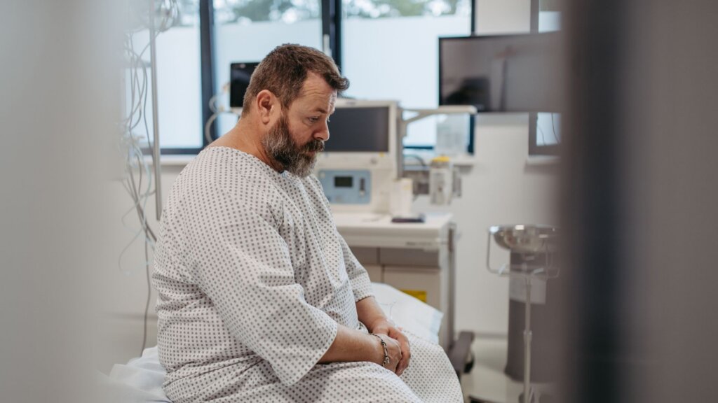 A bearded man in a hospital gown sits on a bed in a modern hospital room, surrounded by medical monitors and equipment.