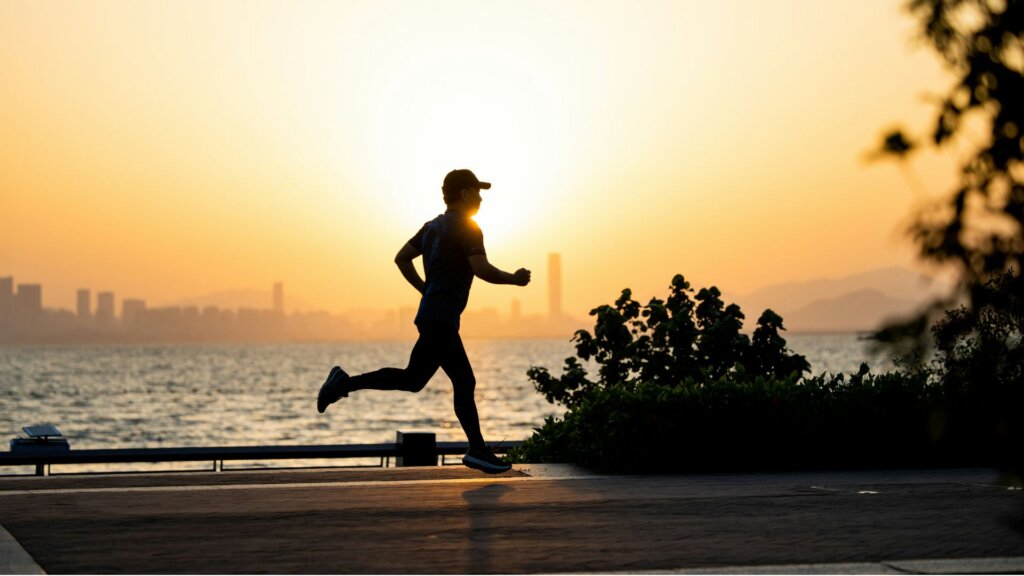 A silhouetted jogger runs along a waterfront at sunset, with a hazy city skyline, trees, and shimmering water.