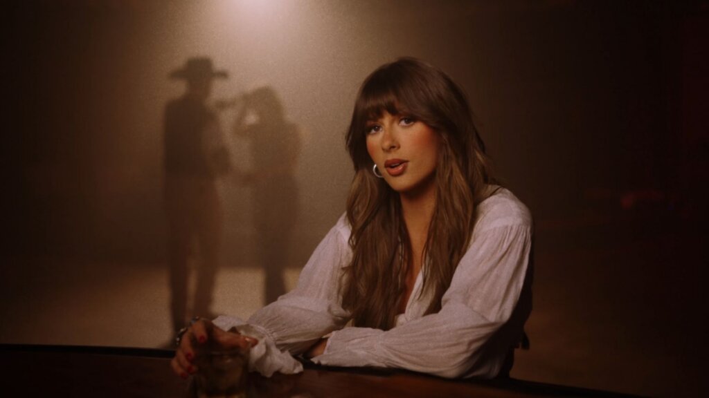 Ella Langley singing at a bar in the 'Choosin' Texas' music video, with figures dancing behind her.