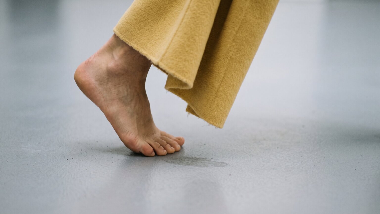A bare foot mid-step on a pale swept floor beside a draping length of butter-yellow wool, standing in for the shoeless Tory Burch look that opens the story.