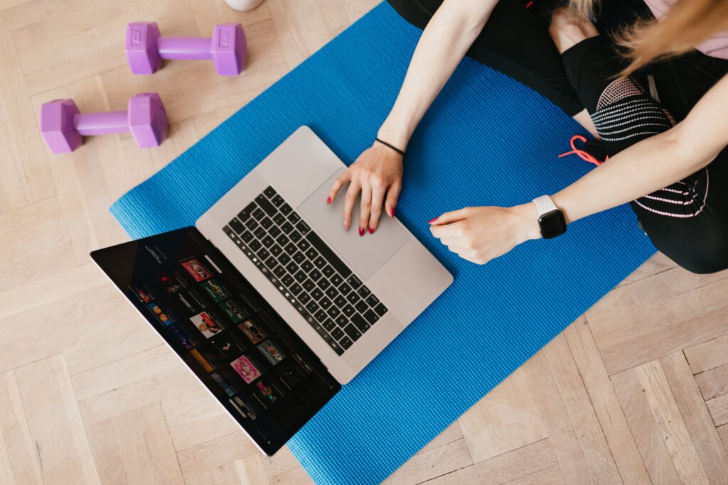 Woman sitting on a blue exercise mat using a laptop, with purple dumbbells nearby, illustrating online fitness training at home.