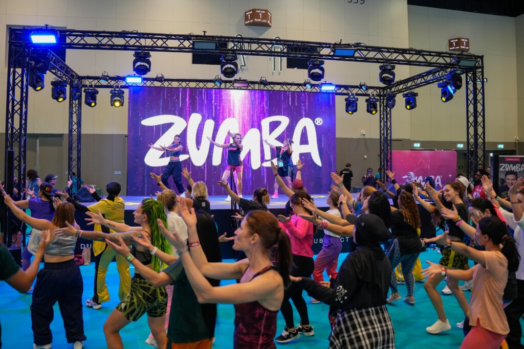 Zumba instructors leading a large dance-fitness class onstage as participants follow their movements beneath a colorful Zumba backdrop.