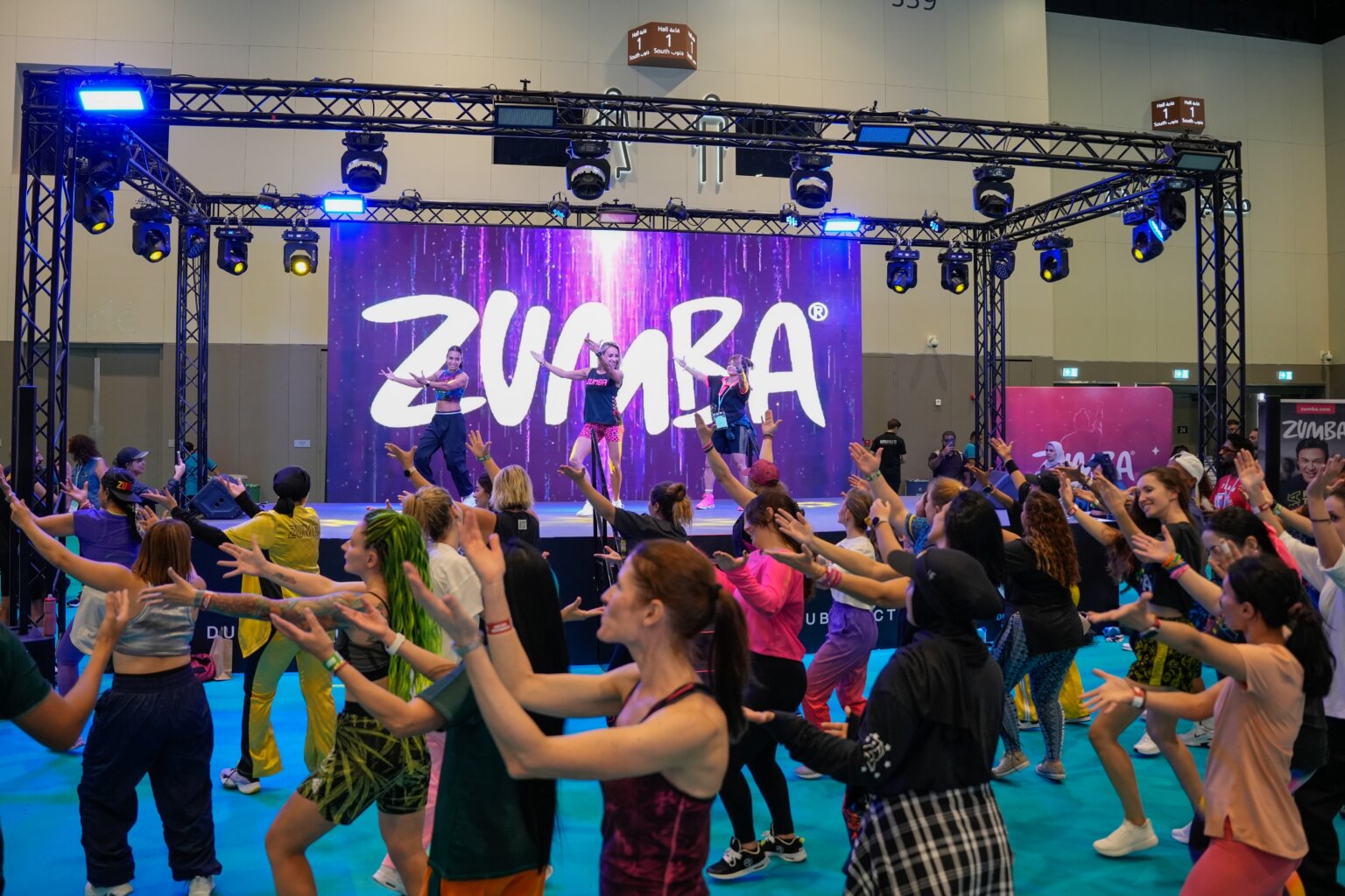 Zumba instructors leading a large dance-fitness class onstage as participants follow their movements beneath a colorful Zumba backdrop.